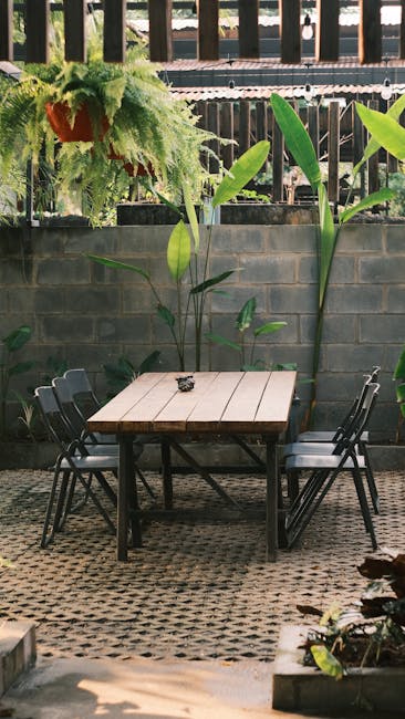 A lush, well-maintained small courtyard garden featuring a mix of vibrant green foliage, including large, broad-leafed plants and neatly trimmed bushes. In the foreground, a round white table with a w